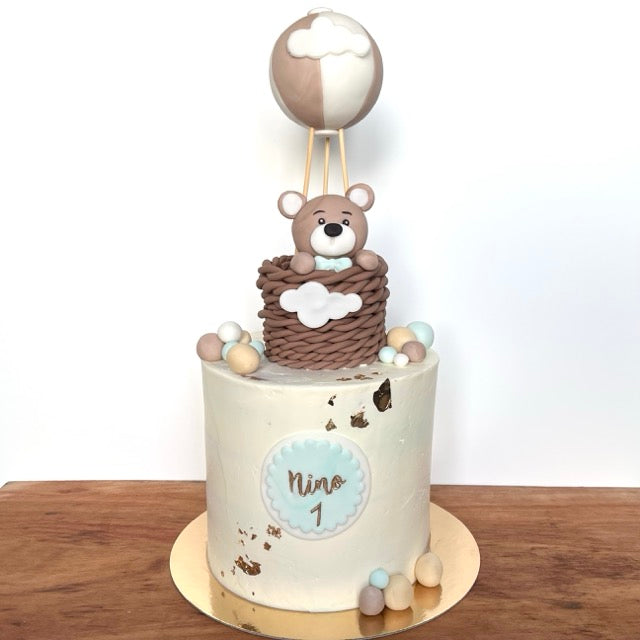 Cake design pour 20 personnes avec un modelage sur le dessus en forme de mongolfière avec un ourson à l'intérieur. Sur le devant du gateau le prénom NINO est inscrit avec le chiffre 1. Pour un gateau d'anniversaire.
