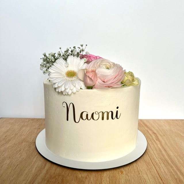 Cake design naked cake avec des fleurs sur le dessus et le prénom naomi écrit sur le devant. 