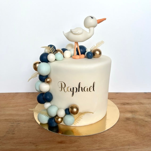 gâteau avec un modelage en pate a sucre avec une cigogne sur le dessus. Des boules bleues marines, bleues ciel, blanches et dorées sur le coté et le prénom Raphaël écrit sur le devant du gâteau.